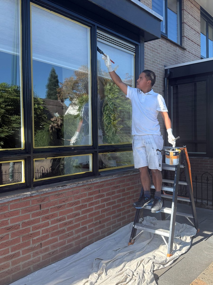Arij lakt een kozijn zwart hoogglans, zijn spiegelbeeld zichtbaar in het glanzende hout